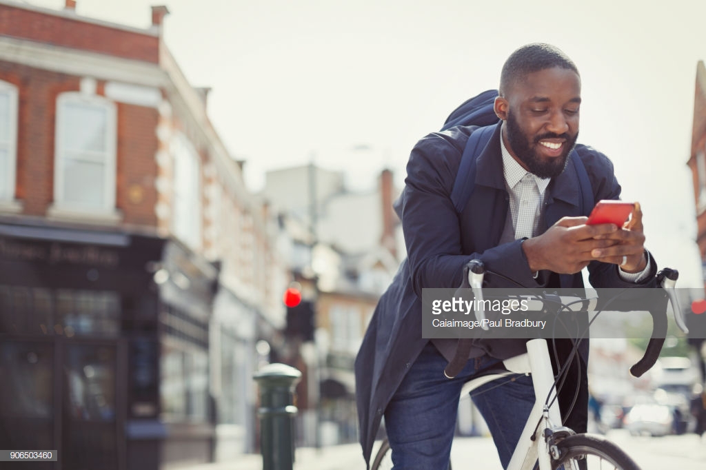 Photo by Paul Bradbury. Young businessman commuting with bicycle, texting with cell phone on sunny urban street.