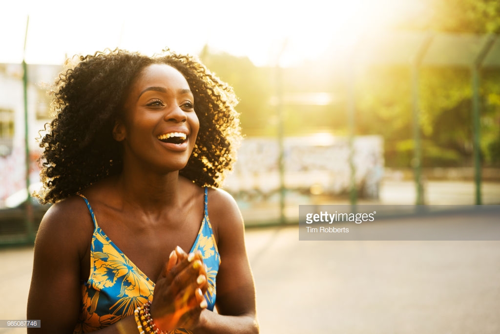 Photograph by Tim Robberts. Close-up of a Black woman smiling, outside on a bright sunny day.