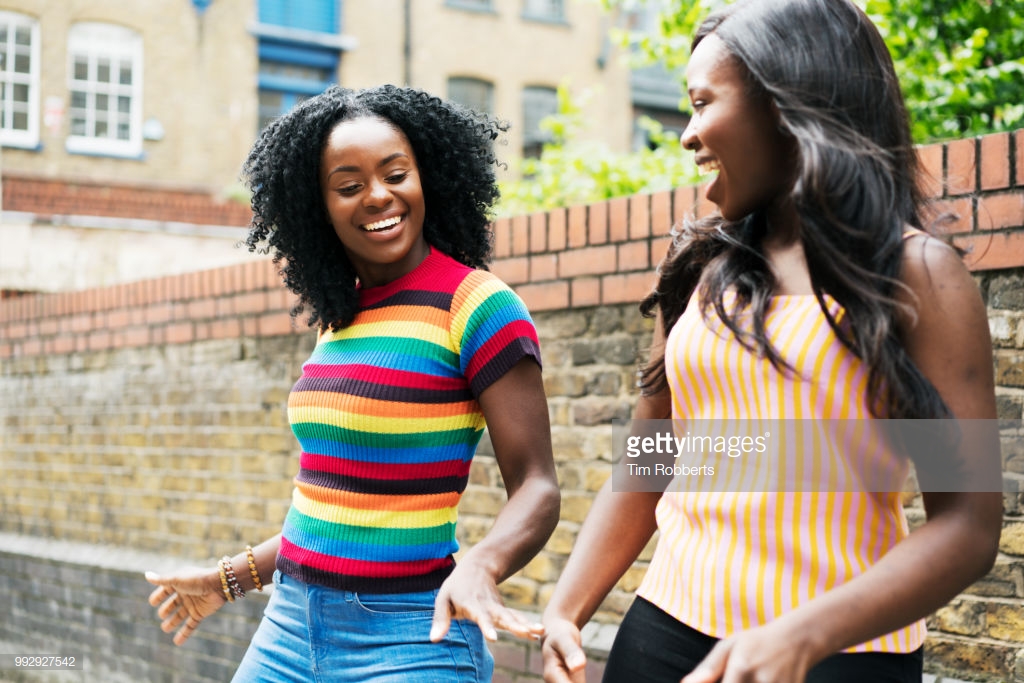 Photograph by Tim Robberts. Two dark-skinned Black women smile together, outside on a bright sunny day.