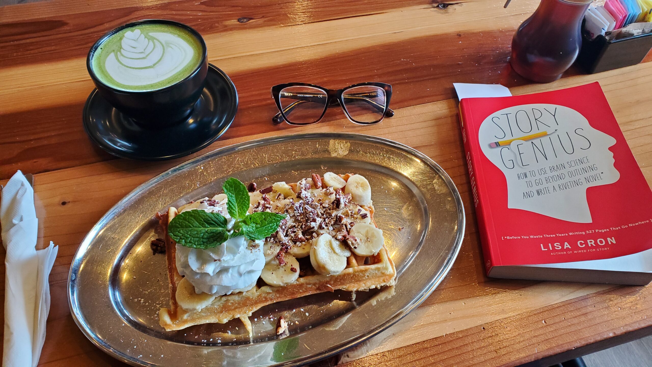 Photo of a meal on a wooden table. A waffle sits on an ovular metal plate. The waffle is topped with bananas, crushed walnuts, whipped cream, and a sprig of mint. Beside the plate is a book: STORY GENIUS by Lisa Cron. Above the plate is a mug of hot matcha latte with a leaf design in cream, and a pair of glasses.