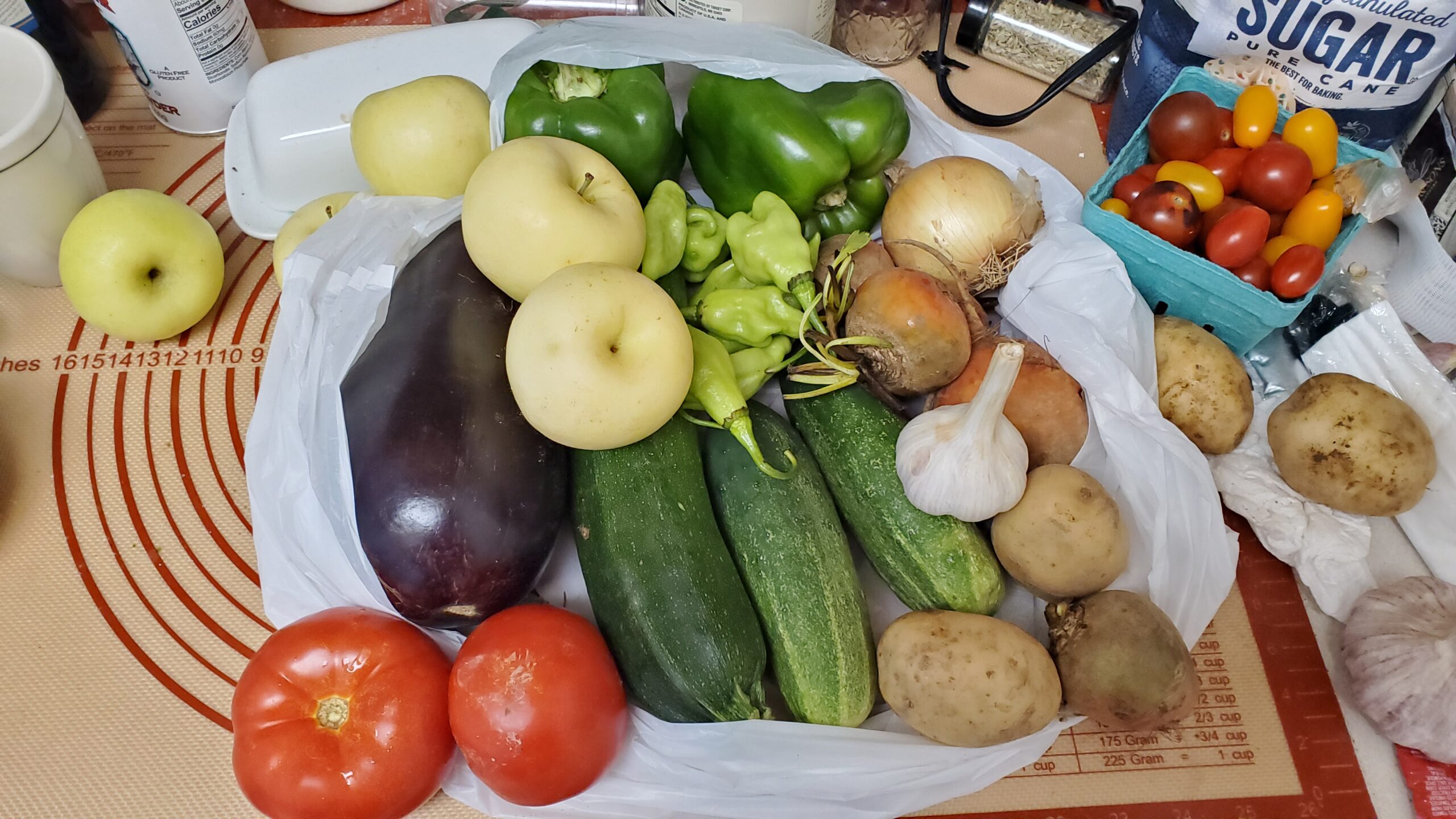 Photo of produce spilling out of a plastic bag on a kitchen countertop: tomatoes, eggplant, apples, peppers, zucchini, tomatoes, potatoes.